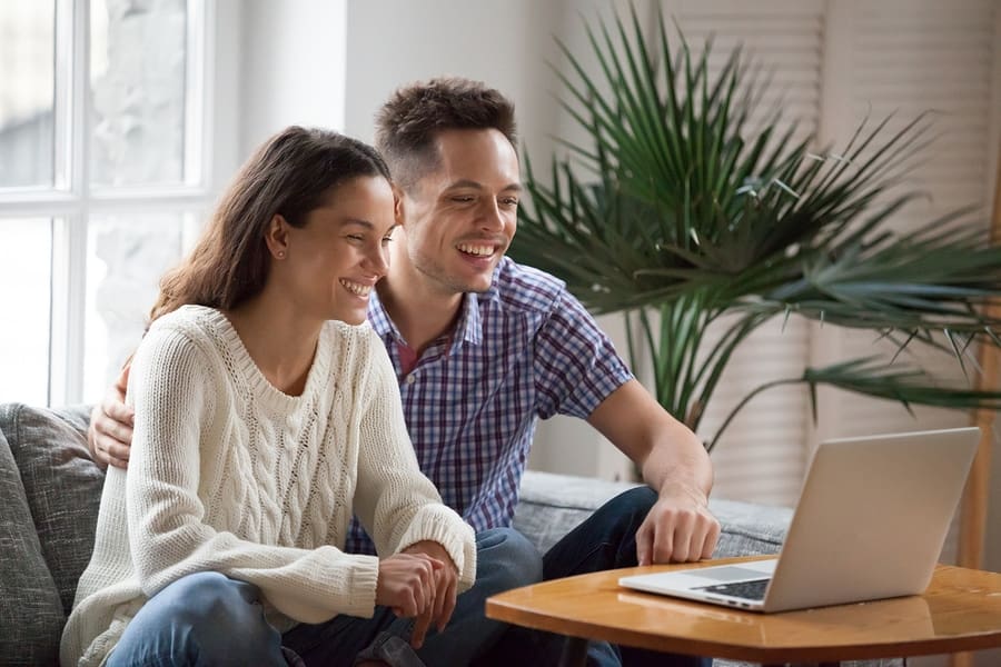 young male and female couple sitting on couch smiling at laptop