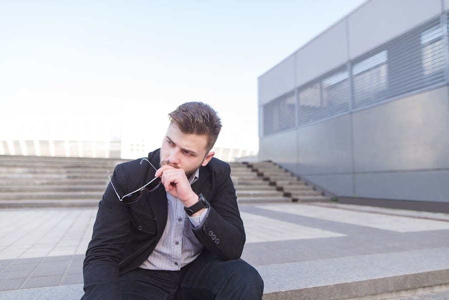 male sitting outside of a building holding glasses looking pensive