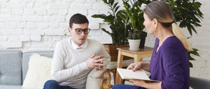 male with glasses on couch talking to female professional with notebook