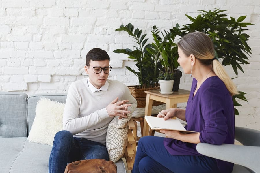 male with glasses on couch talking to female professional with notebook