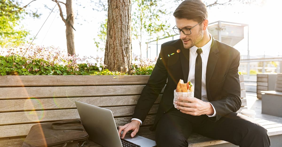 businessman-working-park-bench-laptop-holding-sandwich
