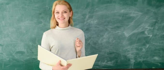Portrait of young teacher in classroom. Young satisfied professor. Prof with folder. Portrait of female teacher with notebooks near chalkboard. Teachers day. Portrait of woman teacher on chalkboard.