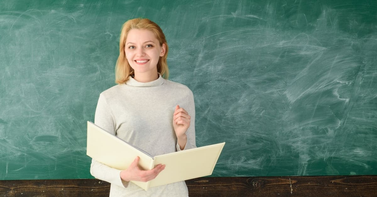Portrait of young teacher in classroom. Young satisfied professor. Prof with folder. Portrait of female teacher with notebooks near chalkboard. Teachers day. Portrait of woman teacher on chalkboard.