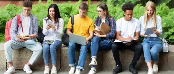 Row of Students Sitting Outside Looking at Books and Devices