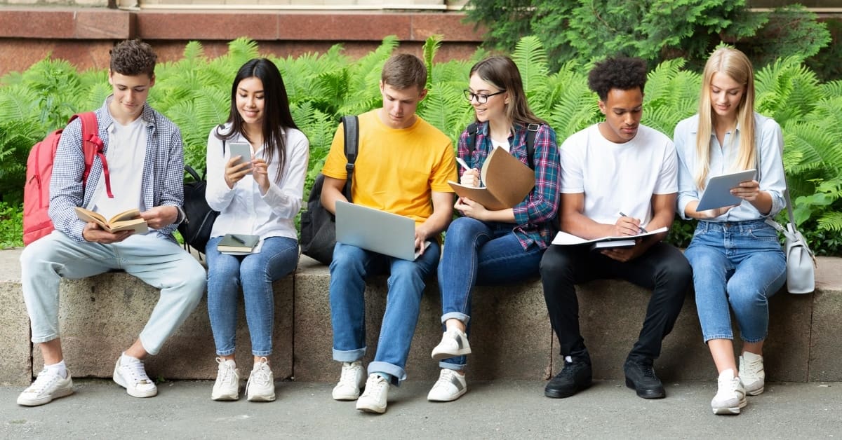 Row of Students Sitting Outside Looking at Books and Devices