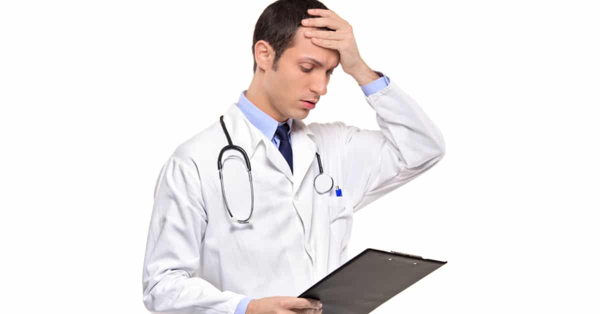 Male Physician with Hand on Forehead Looking at Clipboard