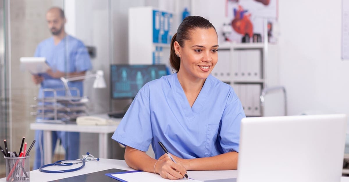 Nurse Smiling Taking Notes While Looking at Laptop