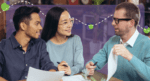 couple having a meeting with male consultant who is holding paper, all sitting on table, all smiling