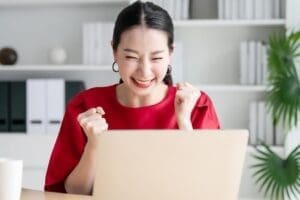 Asian Woman in Red Shirt Celebrating in Front of Laptop