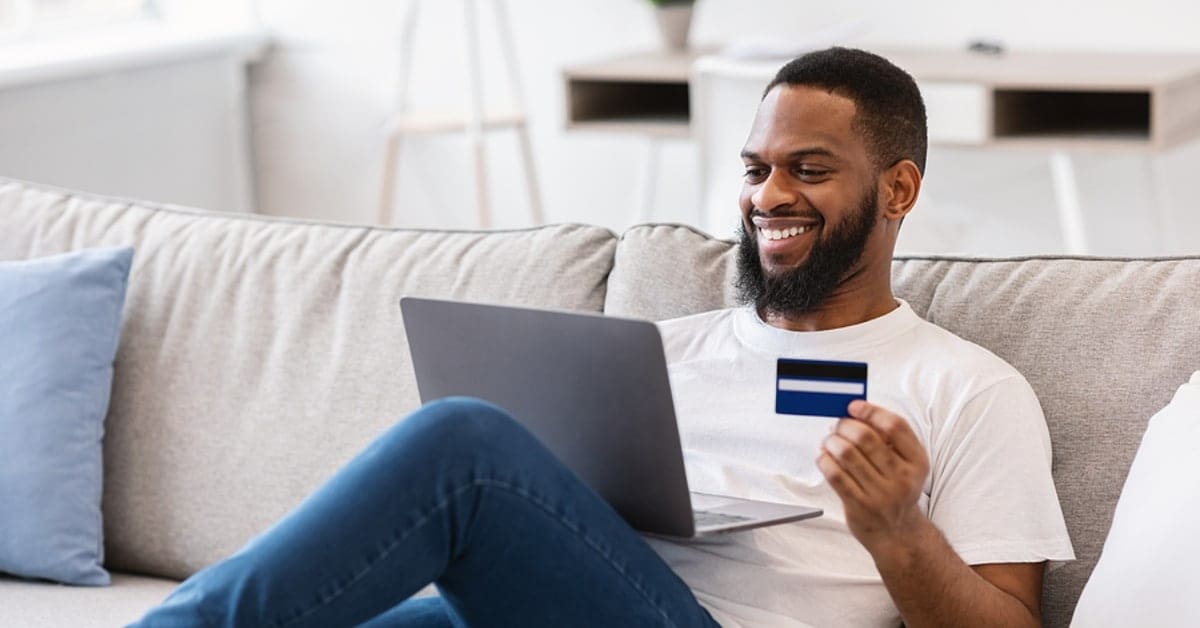 Man Smiling Holding a Credit Card and His Laptop in his Home