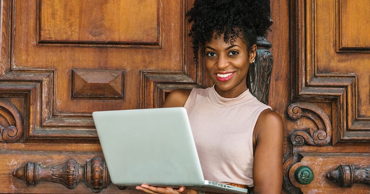 Woman Smiling for Camera While on a Laptop in Front of Large Fancy Wooden Doors