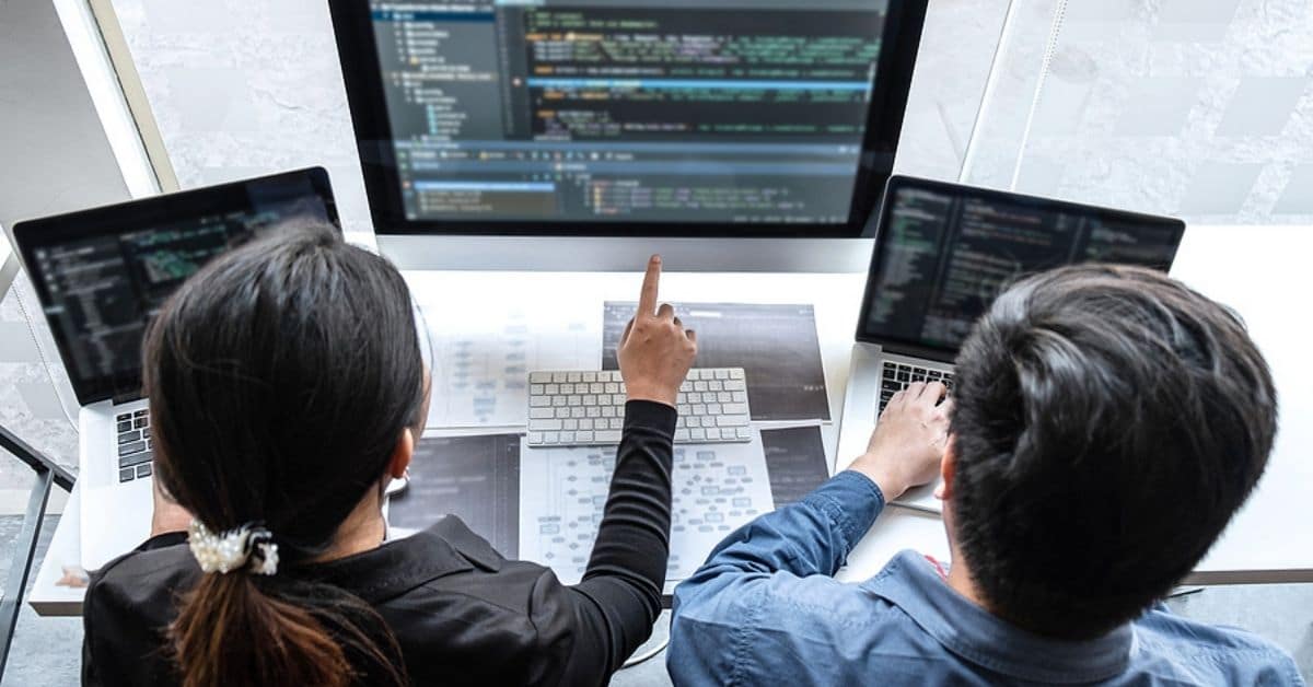 Two Asian Adults Pointing at Three Computers Screens