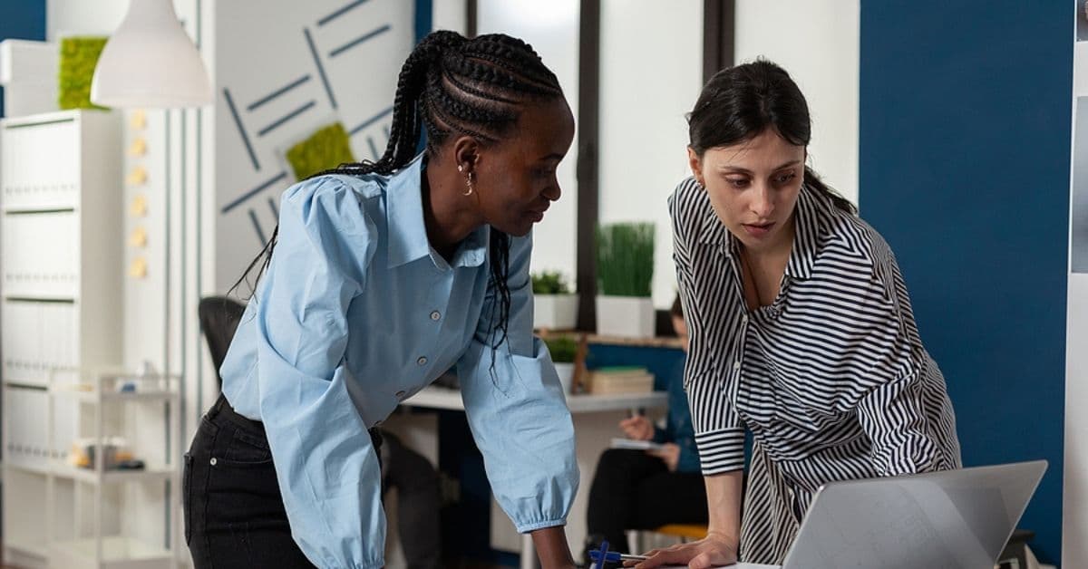 Two Women Leaned Over Discussing Over a Laptop