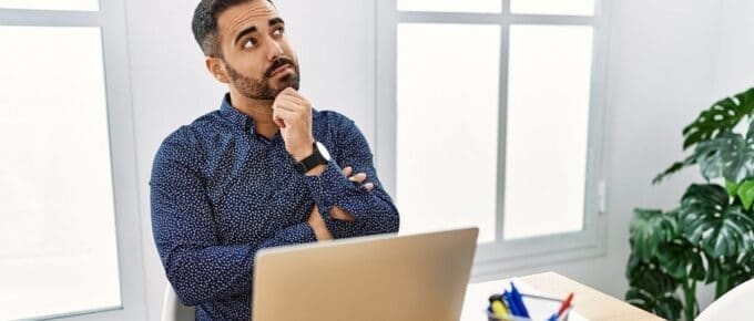 man in blue dress shirt sitting at a desk looking away from laptop