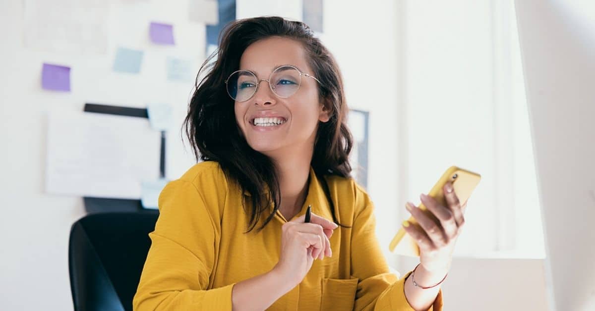 Pretty cheerful businesswoman looking away sitting at desk and holding phone in hand