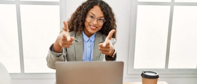 Beautiful hispanic business woman sitting on desk at office working with laptop pointing fingers to camera with happy and funny face.