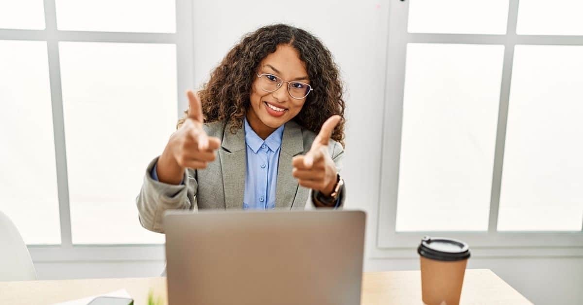 Beautiful hispanic business woman sitting on desk at office working with laptop pointing fingers to camera with happy and funny face.