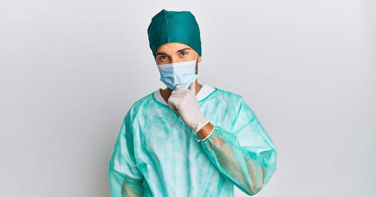 Young handsome man wearing surgeon uniform and medical mask looking confident at the camera with smile with crossed arms and hand raised on chin