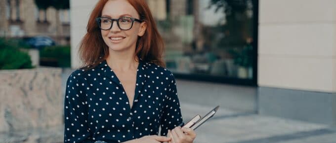 Happy smiling red-haired business woman holding laptop and notebook, looking aside while standing on city street outdoors. Female business professional with digital device near office building
