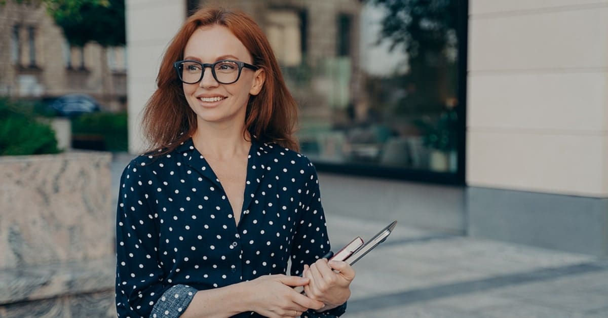 Happy smiling red-haired business woman holding laptop and notebook, looking aside while standing on city street outdoors. Female business professional with digital device near office building
