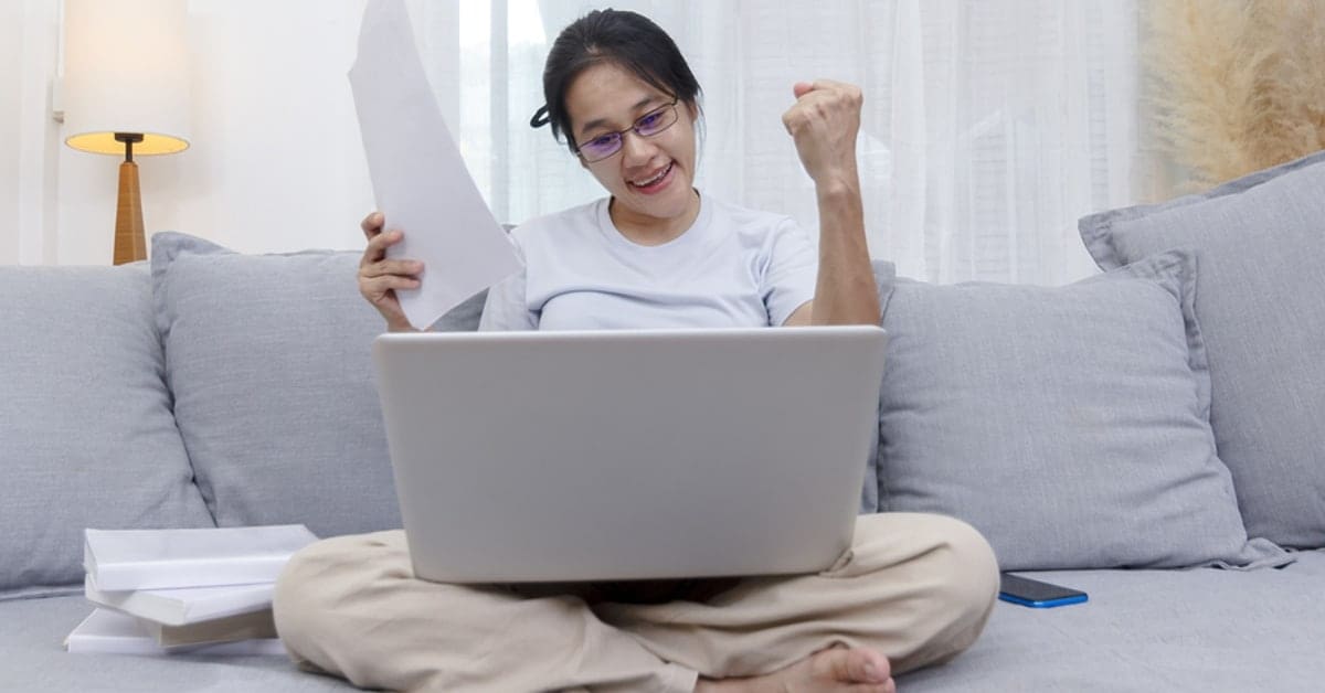 Asian woman celebrating online win success looking laptop at home. Work from home business women sitting on sofa in living room working with laptop online. Young women excited and glad of success with laptop on sofa