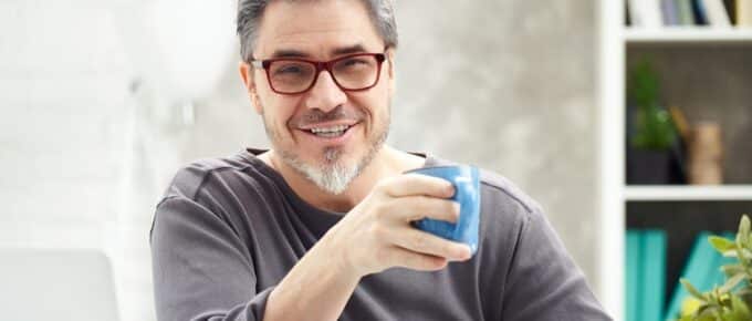 Portrait of happy man at home sitting at desk, working, looking at camera. Happy smile, grey hair, beard, glasses. Portrait of mature age, middle age, mid adult man in 50s.