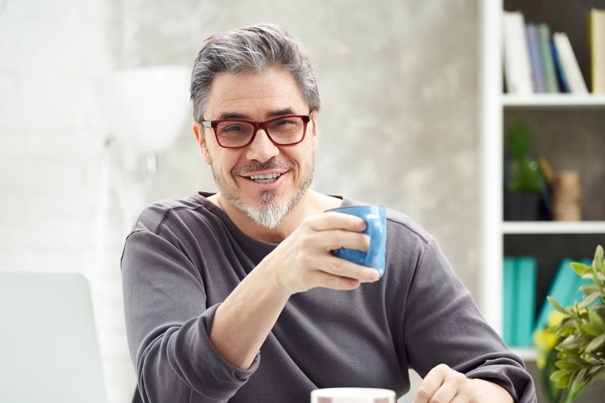 Portrait of happy man at home sitting at desk, working, looking at camera. Happy smile, grey hair, beard, glasses. Portrait of mature age, middle age, mid adult man in 50s.