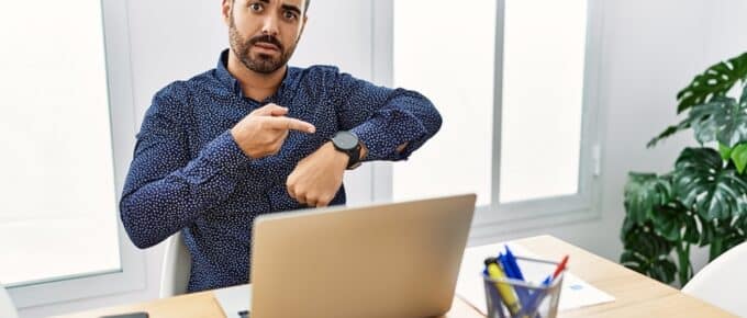Young hispanic man with beard working at the office with laptop in hurry pointing to watch time