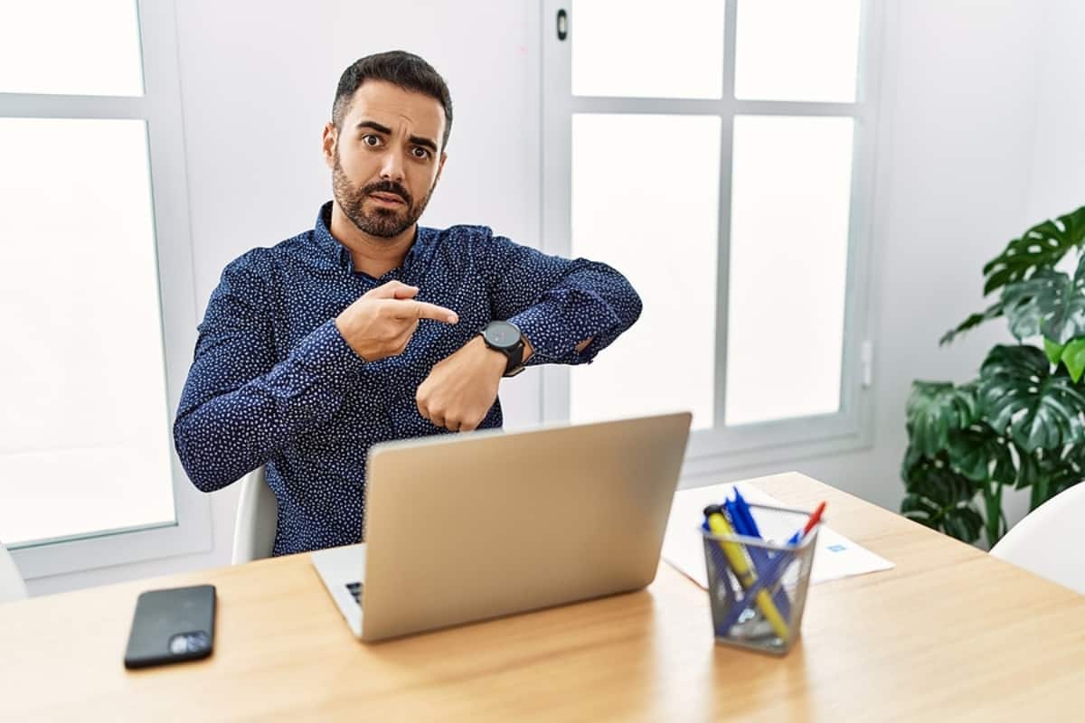 Young hispanic man with beard working at the office with laptop in hurry pointing to watch time