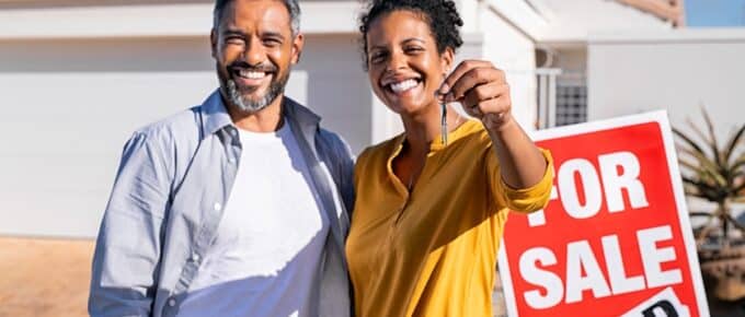 Portrait of happy mixed race couple holding house keys standing near sold signboard. Middle eastern man embracing african woman while showing house key outside of their new home. Moving home day.