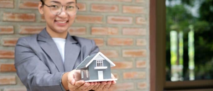 Businesswoman holding a house model in front of a red brick wall.