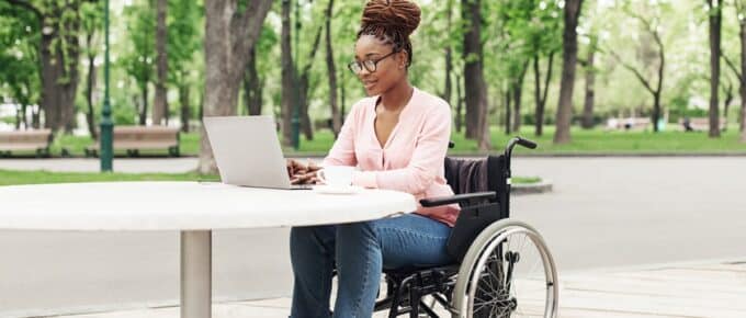 Young black woman in wheelchair working online, using laptop at outdoor cafe