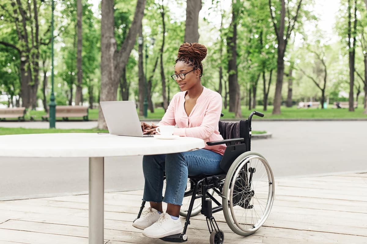 Young black woman in wheelchair working online, using laptop at outdoor cafe