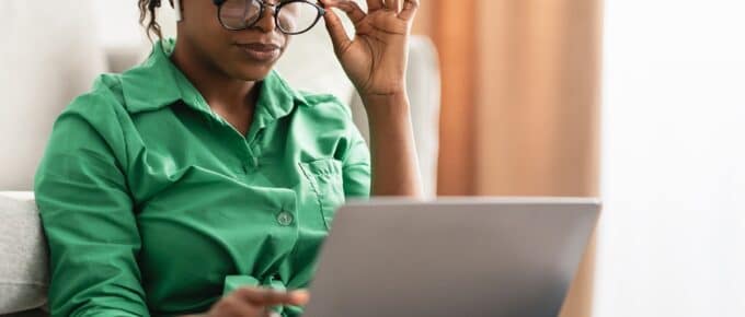Black woman in a green shirt wearing eyeglasses and looking at laptop while sitting on a couch.