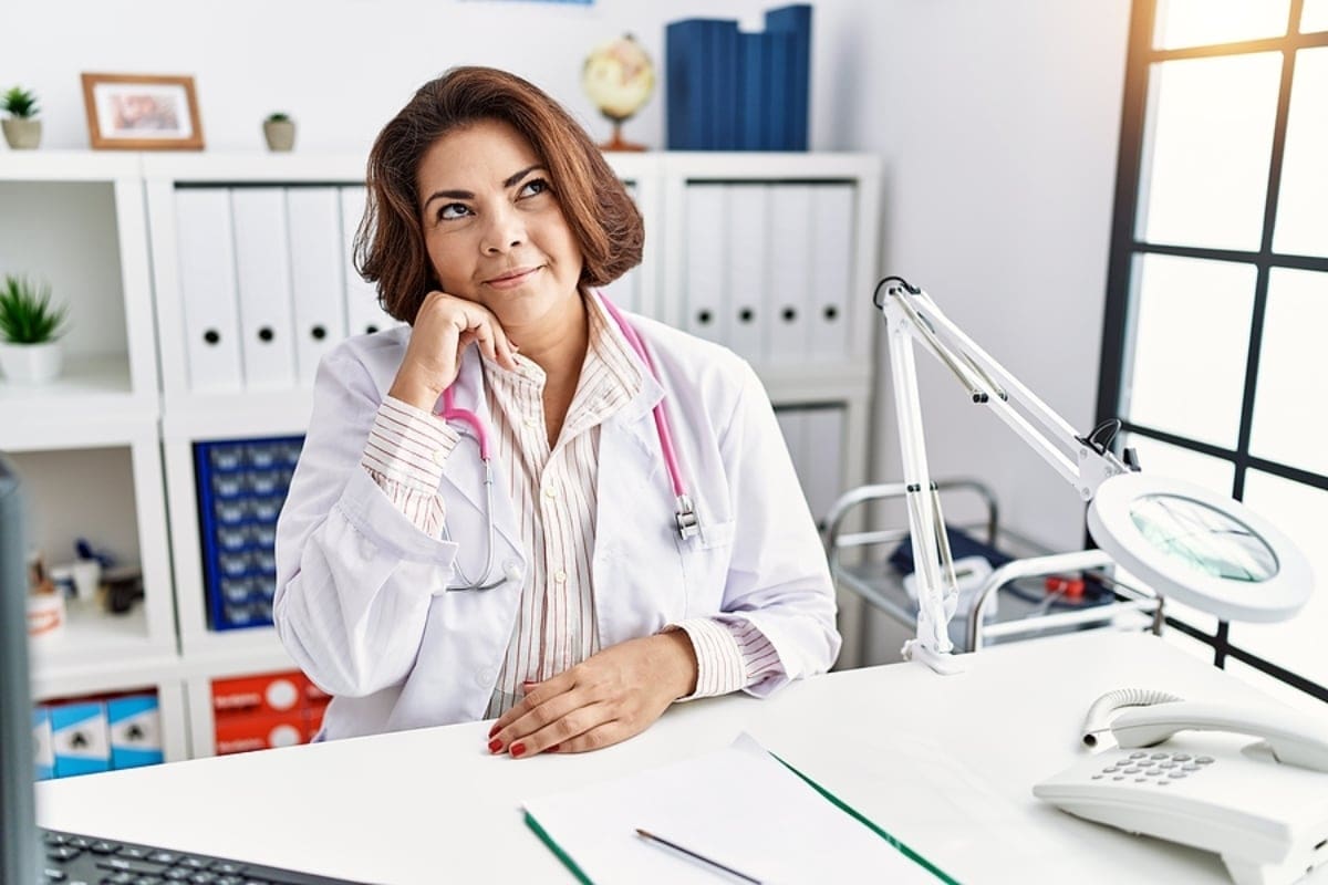 Middle age hispanic woman in doctor uniform and stethoscope at the clinic with hand on chin thinking about question