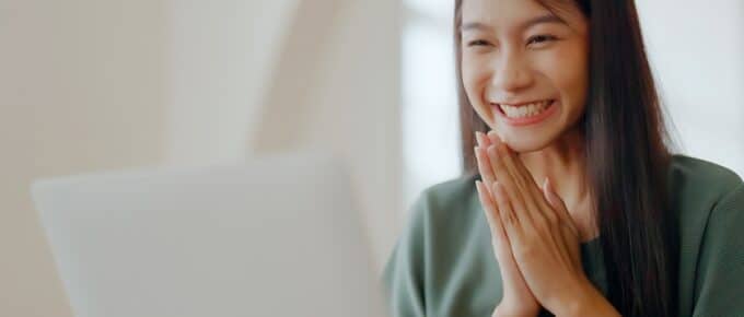 Excited asian female looking at laptop while sitting at a desk in front of a beige wall