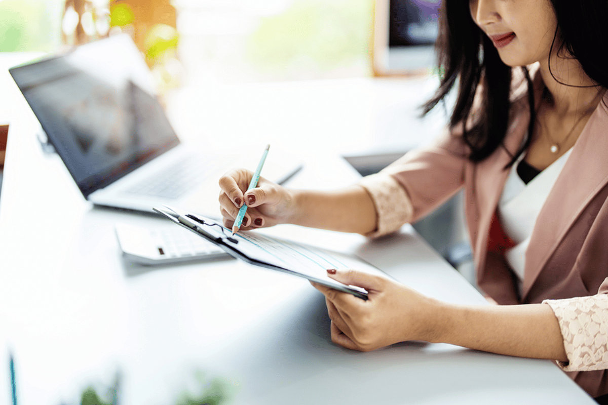 woman with dark hair sitting at a desk holding a clipboard with a laptop in the background