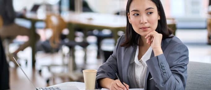 Woman with dark hair wearing a business jacket sitting at a table with a laptop and a coffee thinking.