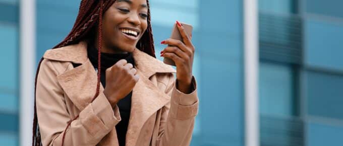 woman in tan coat holding smart phone, looking at it smiling outside in front of a blue building.