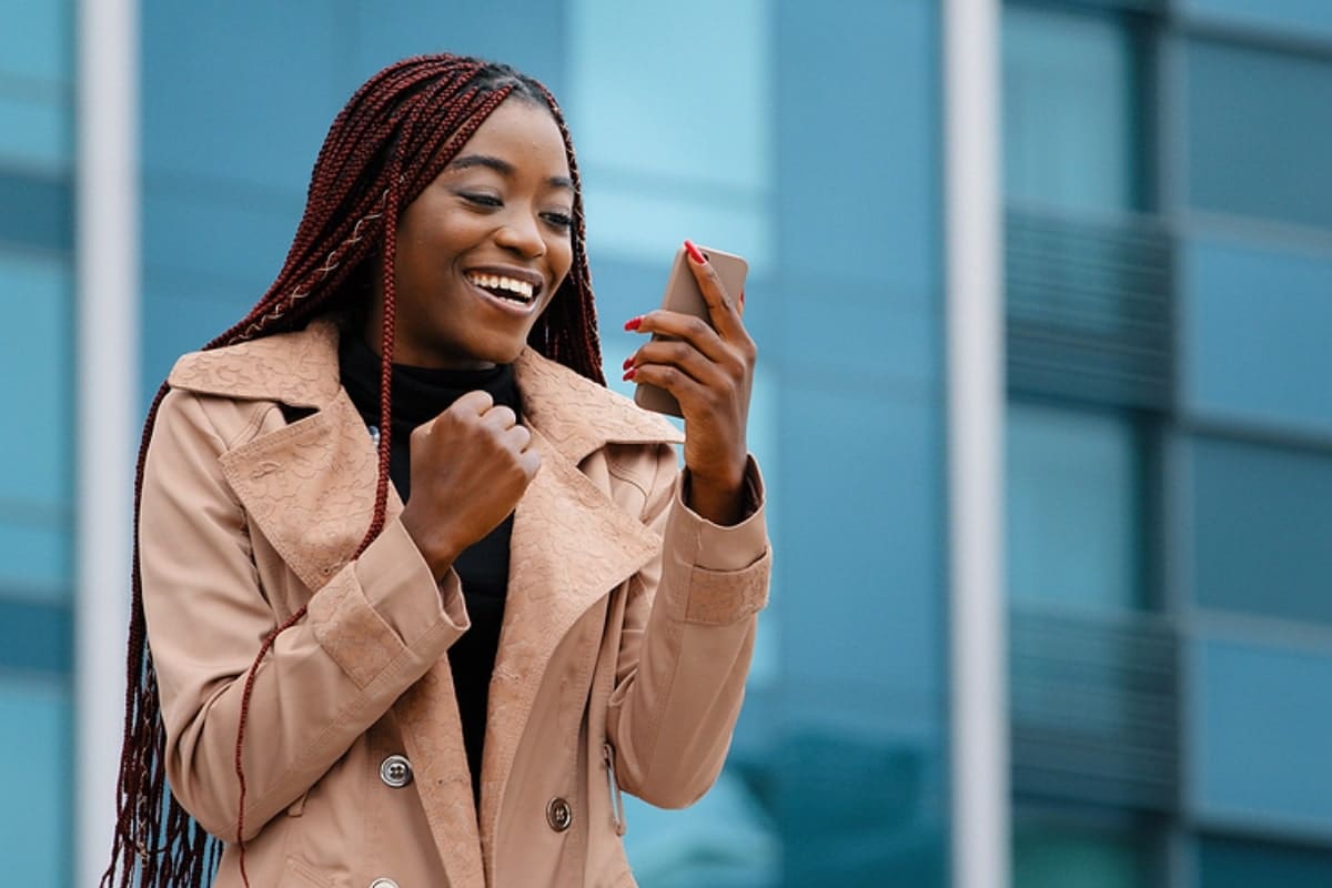 woman in tan coat holding smart phone, looking at it smiling outside in front of a blue building.