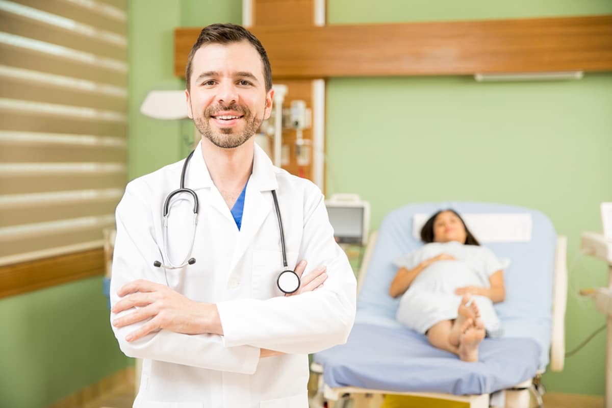 Doctor wearing white coat standing in hospital room with pregnant woman lying in hospital bed in the background.