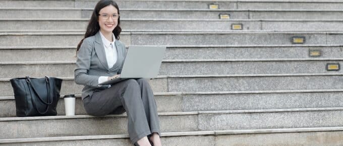 young female entrepreneur sitting on steps next to her bag and cup of coffee and working on laptop