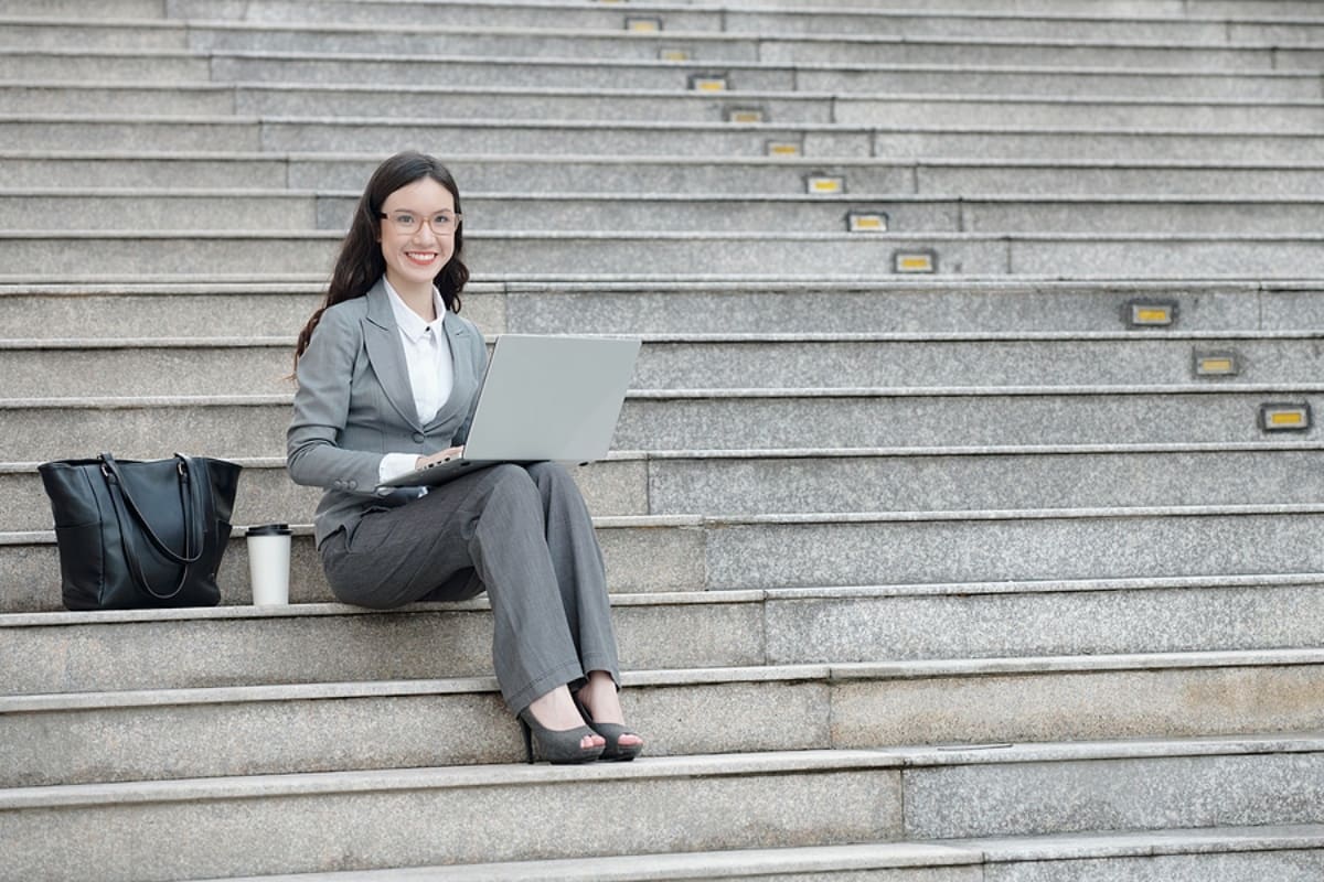 young female entrepreneur sitting on steps next to her bag and cup of coffee and working on laptop