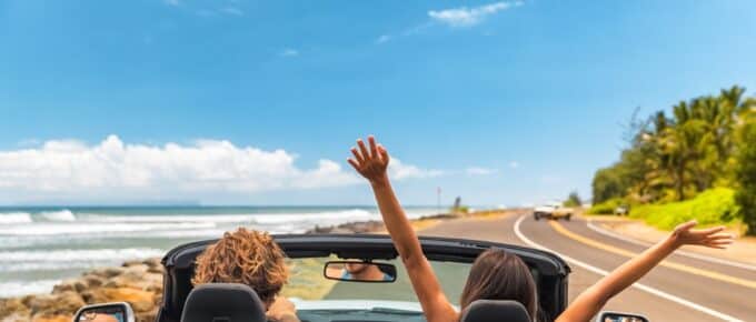 couple in a convertible driving down road in Hawaii with blue skies and the ocean