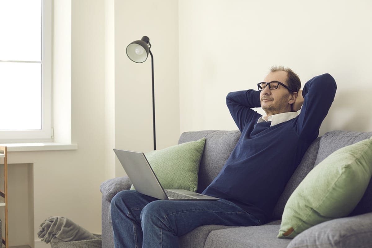 Man took a break from work sitting on a sofa with a laptop and putting his hands behind his head.