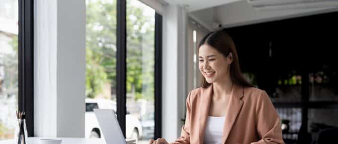 Smiling Asian woman wearing a pink sweater, sitting at a desk, using a laptop