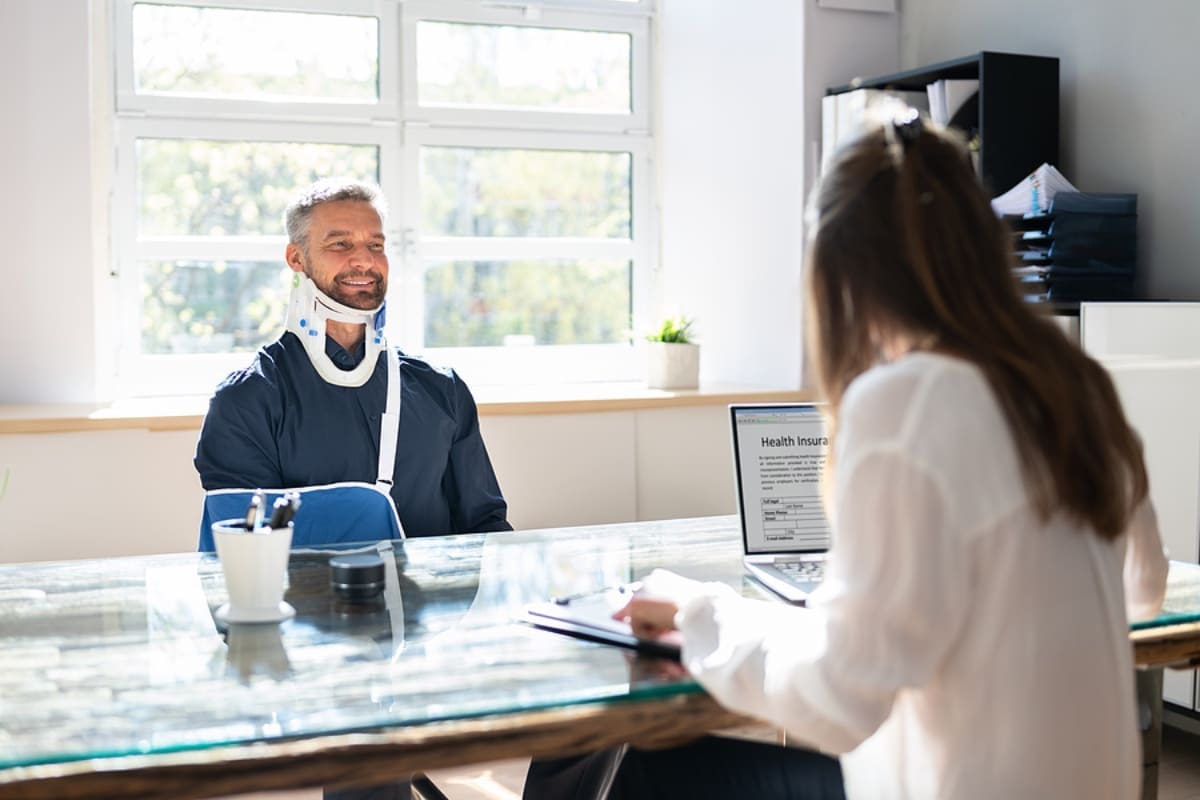 Man wearing blue shirt sitting in wheel chair with neck brace talking to a dark haired woman sitting behind a desk filling out disability insurance forms.