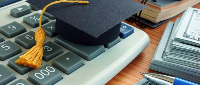 Calculator with piles of money and small graduation cap sitting on a desk.