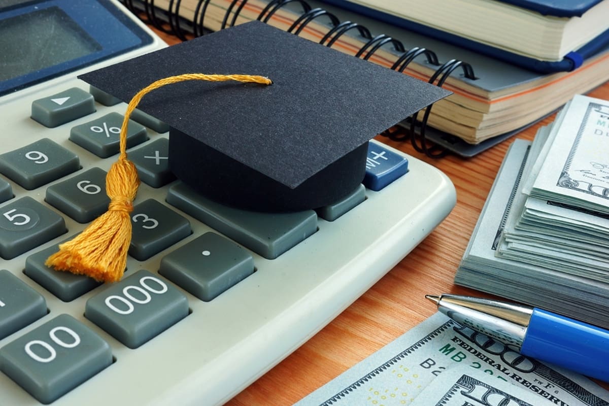 Calculator with piles of money and small graduation cap sitting on a desk.