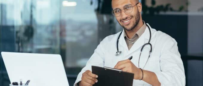 Male doctor smiling and looking at camera, using laptop for work.
